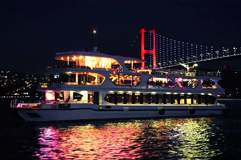 یک کشتی تفریحی شبانه در بسفر، در حال عبور از مقابل یک پل چراغانی